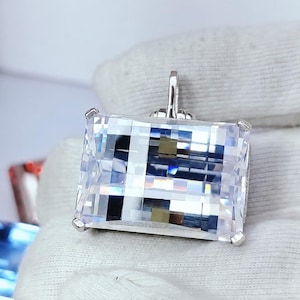 Pode incluir: Um grande pingente de pedra preciosa transparente, retangular e facetada, montado em uma armação de metal prateado com garras. A pedra apresenta um corte brilhante com inúmeras superfícies reflexivas que criam um padrão de luz.
