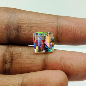 Puede incluir: Una gema de corte cuadrado con un interior multicolor vibrante. La gema muestra un espectro de colores, incluyendo rojo, amarillo, verde y azul, creando un efecto deslumbrante. La gema se sostiene entre los dedos, mostrando su tamaño y claridad.