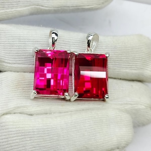 May include: Two rectangular, faceted, hot pink gemstone pendants with silver settings. Each pendant has a loop at the top for a chain. The gems are a vibrant, saturated colour, and the setting is a bright, shiny metal.