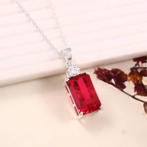 May include: A silver necklace with a rectangular, bright red gemstone pendant. The pendant has a smaller, round, clear stone above the main gem. The necklace chain is delicate and silver.
