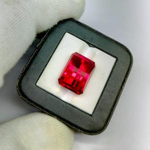 Puede incluir: Una vibrante gema roja, de corte rectangular, exhibida en una caja de presentación en blanco y negro. Las facetas de la gema reflejan la luz, mostrando su claridad y color. La caja es sostenida por una mano enguantada.