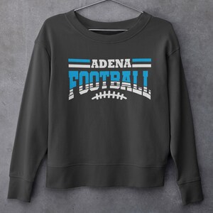 May include: Dark gray sweatshirt with the text "ADENA FOOTBALL" in white and blue. The word "FOOTBALL" is stylized with a football graphic. The sweatshirt is hanging on a metal hanger against a gray wall.