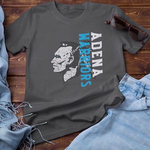 May include: Dark gray t-shirt featuring a side profile of a Native American chief with the text "ADENA WARRIORS" in white and blue. Sunglasses and denim jeans are also visible.