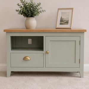 May include: A sage green media console featuring a natural wood top, one open shelf, one drawer with a gold half-moon pull, and one cabinet door with a gold round knob. A potted green plant and a framed landscape print sit on the wooden surface.
