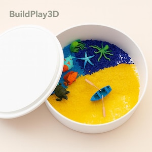 May include: A white container with a lid, filled with blue and yellow textured material, resembling water and sand. Miniature sea creatures, including a starfish, octopus, and turtle, are scattered throughout. A small blue boat with oars is also present. The text "BuildPlay3D" is visible.