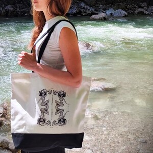 May include: A white canvas tote bag with black handles and a black bottom. The bag has a black and white graphic print of two women in a symmetrical design.