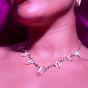 May include: A gold-toned metal necklace featuring a series of evenly spaced cowrie shell-shaped charms linked by a delicate chain, accompanied by a matching single cowrie shell-shaped earring hanging from a thin metal hook.