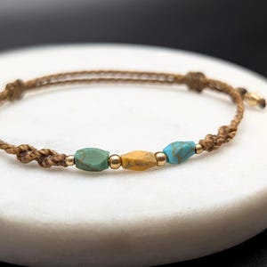 Puede incluir: Un brazalete de cordón marrón con tres cuentas: una azul turquesa, una amarilla y otra azul turquesa. El brazalete está sobre una superficie blanca.