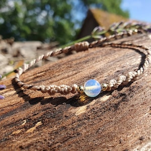 May include: A light blue opal bead on a brown braided cord bracelet. The bracelet is lying on a piece of weathered wood.