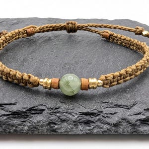 Könnte beinhalten: Ein braunes geflochtenes Armband mit einem grünen Edelsteinperlen und goldenen Akzenten.