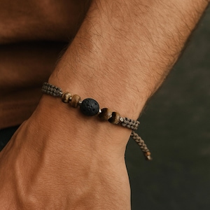 Könnte beinhalten: Ein gewebtes Armband mit einer schwarzen Lavasteinperle, Holzperlen und einer geflochtenen Kordel. Das Armband hat eine neutrale Farbpalette mit Braun- und Beigetönen. Das Armband wird am Handgelenk getragen.