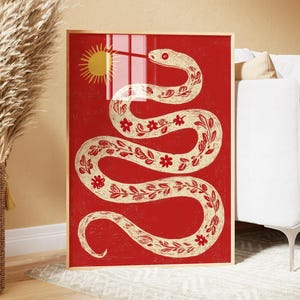 Puede incluir: Una lámina de arte mural rectangular en un marco de madera clara con fondo rojo y una gran serpiente de color crema que se enrosca en forma de S. La serpiente está decorada con pequeñas flores y hojas rojas, y un sol amarillo se sitúa arriba a la izquierda.
