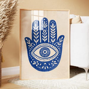 May include: A rectangular framed wall art print featuring a bold blue Hamsa hand illustration centered on a light beige background. The hand design includes intricate white botanical leaf patterns within the fingers and a central eye symbol.