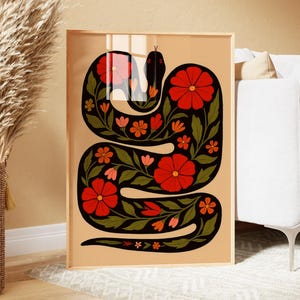 Op de afbeelding: Ingelijste kunstprint met een gestileerd zwart slangenontwerp versierd met rode en oranje bloemen en groene bladeren. Het kunstwerk heeft een beige achtergrond en is ingelijst in een lichte houten lijst. De print heeft een bohemien of retro esthetiek.