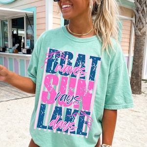 May include: A light green t-shirt with the words "Boat Waves Sun Rays Lake Days" in a vertical design. The text is in blue and pink, with a distressed, vintage style. The shirt has short sleeves and a relaxed fit.