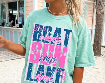Könnte beinhalten: Hellgrünes T-Shirt mit dem vertikalen Schriftzug "Boat Waves Sun Rays Lake Days". Der Text ist in Blau und Pink gehalten, im Vintage-Stil. Das Shirt hat kurze Ärmel und eine lockere Passform.