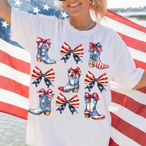 May include: White t-shirt featuring watercolor illustrations of cowboy boots and bows in red, white, and blue, with stars and stripes designs. The boots and bows are arranged in a repeating pattern, evoking a patriotic theme.