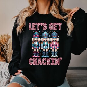 May include: Black sweatshirt with the text "LET'S GET CRACKIN'" in pink glitter lettering. The design features three colourful nutcracker figures in blue, pink, and teal, each wearing a crown. A festive holiday item.