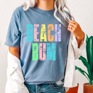 May include: A light blue t-shirt with the text "Beach Bum" in rainbow colors. The text has a distressed, vintage look.