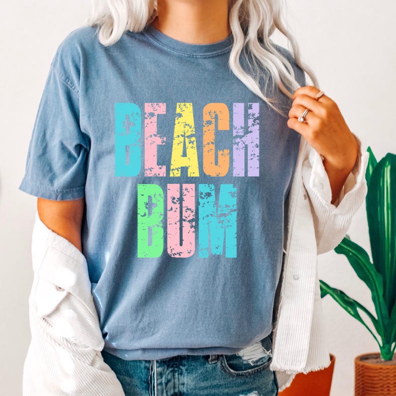 May include: A light blue t-shirt with the text "Beach Bum" in rainbow colors. The text has a distressed, vintage look.