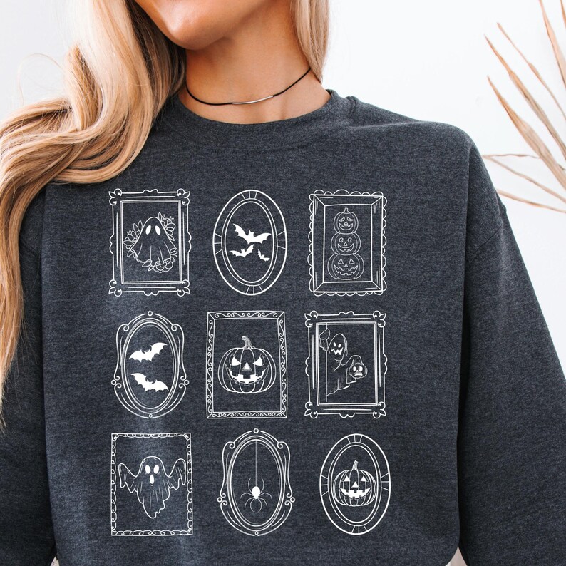 May include: Dark gray sweatshirt featuring nine framed Halloween-themed illustrations in white. The frames contain images of ghosts, bats, pumpkins, and a spider. The sweatshirt has a crew neck and long sleeves.