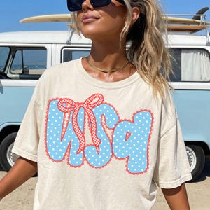 May include: Cream-colored t-shirt with a blue polka-dot design spelling out "USA" with a red bow. The shirt is worn by a person with sunglasses and a gold necklace. A light blue van with a surfboard on top is in the background.