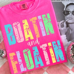 May include: A bright pink t-shirt with the text "BOATIN and FLOATIN" in rainbow colors with a distressed, vintage look.