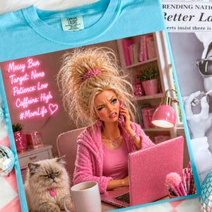 May include: Light blue t-shirt with a graphic print. The print features a doll-like figure with a messy bun, on the phone, and a cat. Text includes "Messy Bun, Target: None, Patience: Low, Caffeine: High, #MomLife".