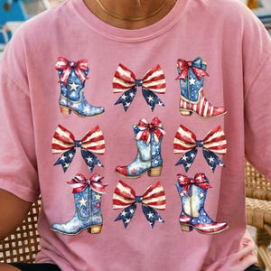 May include: Pink t-shirt featuring a repeating pattern of watercolor cowboy boots and bows. The boots are blue with white stars and red accents, while the bows display a red, white, and blue American flag design.