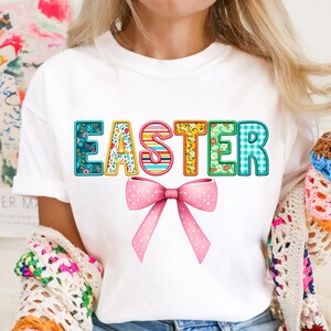 May include: White t-shirt with the word "EASTER" spelled out in colorful patterned letters. A pink bow with white polka dots is centered below the word.