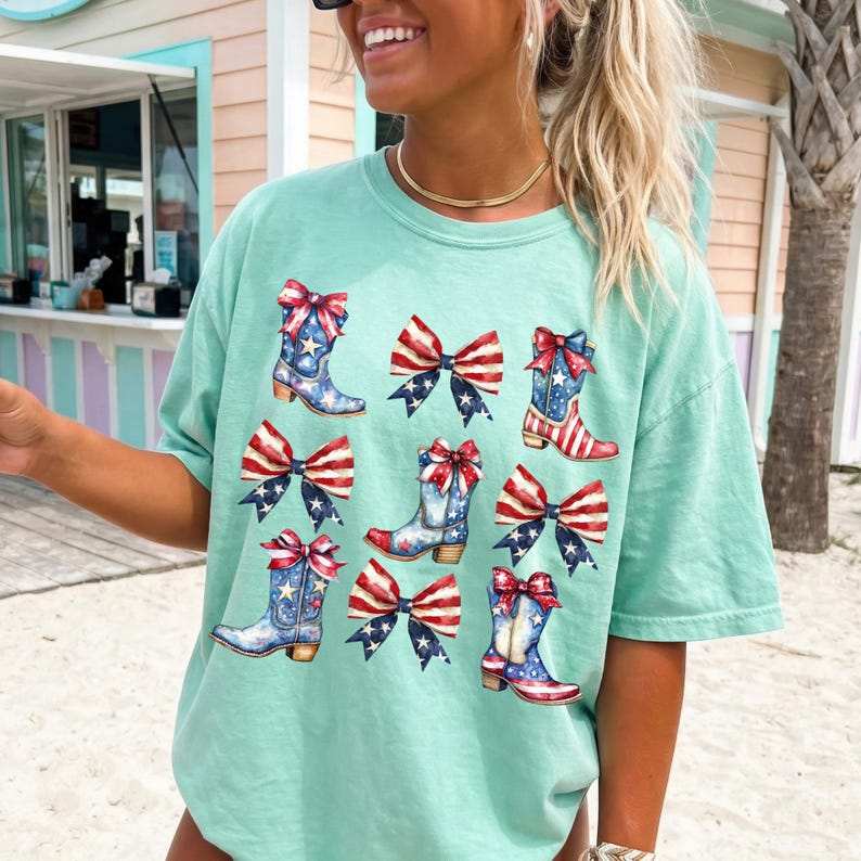 May include: A light green t-shirt featuring a patriotic design. The design includes illustrations of cowboy boots and bows in red, white, and blue, with stars and stripes patterns. The shirt is a casual, comfortable style.