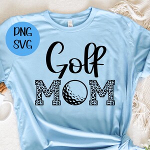 Op de afbeelding: Een lichtblauw T-shirt met de tekst "Golf Mom" in zwart. De letters "M" en "O" zijn versierd met een luipaardprint. Een zwart-witte golfbal bevindt zich in het midden tussen de letters.