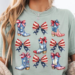 May include: A light green t-shirt featuring a watercolor design of cowboy boots and bows in red, white, and blue. The boots have stars and stripes, and the bows have a patriotic theme.