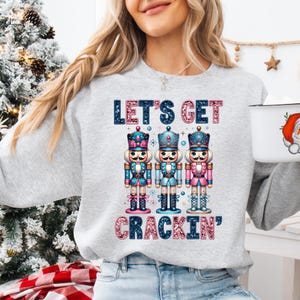 May include: A grey sweatshirt with a festive Christmas design featuring three nutcracker figurines and the text "Let's Get Crackin'" in pink and blue glitter.