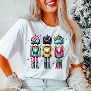 May include: White t-shirt with a graphic of three decorative nutcrackers. The nutcrackers are in bright colours, including pink, blue, green, and gold. Each has a unique hat and design, perfect for the festive season.