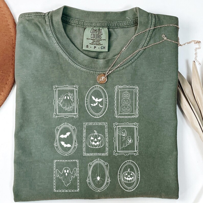 May include: Olive green Comfort Colors t-shirt with a white framed Halloween-themed graphic. The graphic includes bats, ghosts, pumpkins, and spiders. A gold necklace with a pendant rests on the shirt.