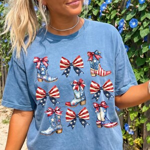May include: A light blue t-shirt featuring a patriotic design. The design includes illustrations of cowboy boots and bows, decorated with red, white, and blue stars and stripes. The shirt has a relaxed fit.