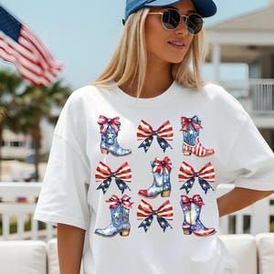 May include: White t-shirt featuring a patriotic design of cowboy boots and bows in red, white, and blue. The boots have stars and stripes, and the bows have a similar pattern. The design is repeated across the shirt.