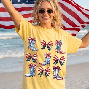 May include: Yellow t-shirt featuring a patriotic design with watercolor illustrations of cowboy boots and bows in red, white, and blue. The boots have star and stripe patterns. The shirt is worn by a person holding an American flag.