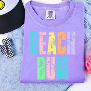 May include: A purple t-shirt with the words "Beach Bum" in rainbow colors. The shirt has a distressed look and is made by Comfort Colors.