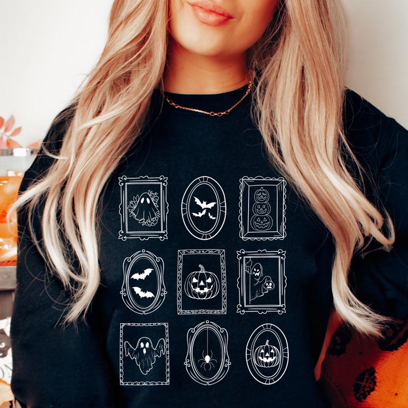 May include: Black sweatshirt featuring a white Halloween-themed design. The design includes framed illustrations of ghosts, bats, pumpkins, and spiders. The frames are in various shapes, including rectangular and oval.