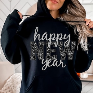 以下が含まれることがあります： 「happy new year」と銀色のグリッターデザインが施された黒いパーカーです。