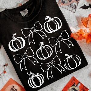 Puede incluir: Camiseta negra con un diseño gráfico blanco de calabazas y lazos. El diseño se repite en la parte delantera de la camiseta. La camiseta está doblada sobre una superficie clara, con decoraciones de Halloween en el fondo.