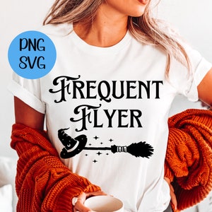 以下が含まれることがあります： 黒い筆記体で「Frequent Flyer」と書かれた白いTシャツ。黒い魔女の帽子とほうきがテキストの上に飛んでいます。
