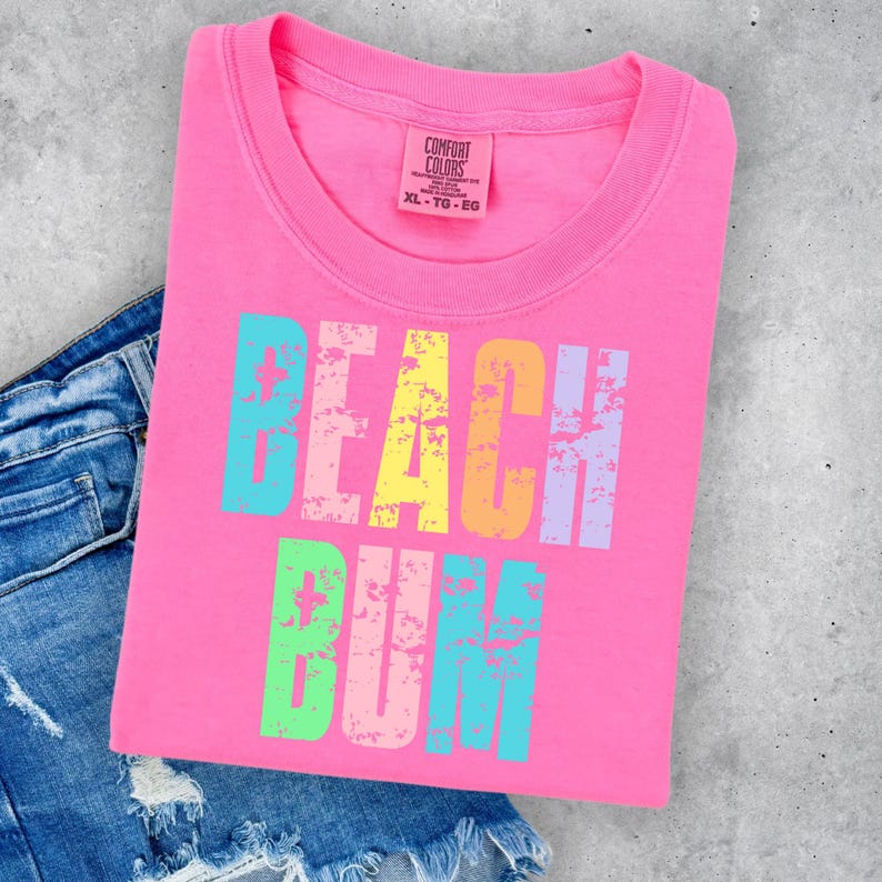May include: Pink t-shirt with the text "Beach Bum" in rainbow colors with a distressed look. The shirt is folded and laying on a pair of blue jeans.