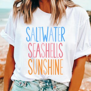以下が含まれることがあります： レインボーグラデーションで「Saltwater Seashells Sunshine」と書かれた白いTシャツです。