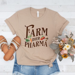 May include: A tan t-shirt with the text "FARM OVER PHARMA" in brown lettering. The text is surrounded by small images of vegetables and a mushroom.