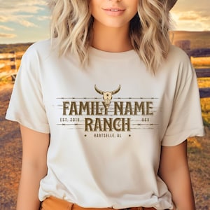 Puede incluir: Una camiseta blanca con un diseño gráfico marrón que dice "FAMILY NAME RANCH" con un logotipo de calavera y huesos cruzados. El texto también dice "EST. 2018" y "HARTselle, AL".
