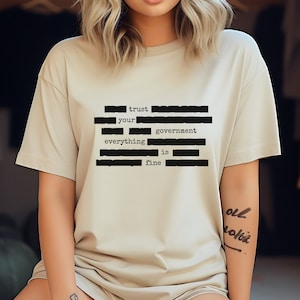 Può includere: Una t-shirt beige con una grafica nera che dice "trust your government everything is fine".