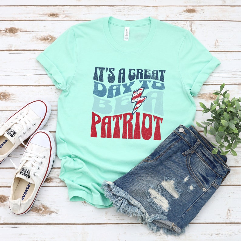 May include: A light blue t-shirt with a distressed graphic that reads "It's a great day to be a patriot". The graphic features a red, white and blue lightning bolt.